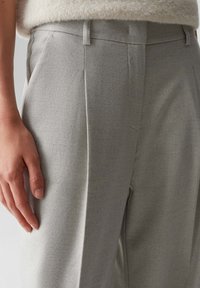 Un pantalon gris sur mesure en tissu texturé, avec une taille haute, des passants de ceinture et un pli marqué à l'avant. Main reposant sur le tissu.
