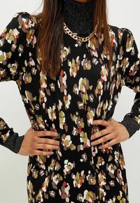 Robe noire à motifs floraux avec des manches longues. Équipée d'un col roulé au look métallique et d'un collier en chaîne dorée. Texture douce et coupe décontractée.