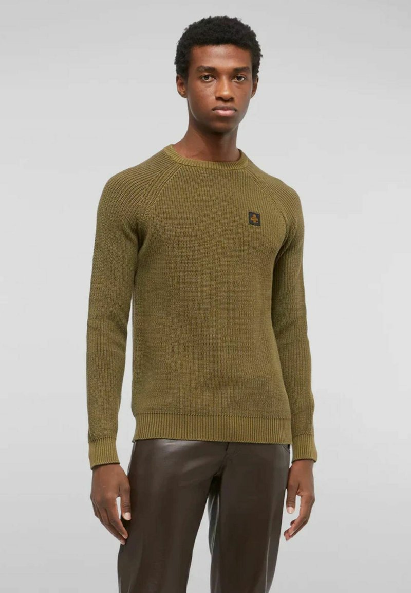 Uomo che indossa un maglione lavorato a maglia verde oliva e pantaloni in pelle marrone scuro, in piedi contro uno sfondo grigio chiaro uniforme.