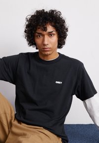 Μαύρη βαμβακερή μπλούζα T-shirt με λευκό λογότυπο "OBEY" μπροστά, συνδυασμένη με ανοιχτό καφέ παντελόνι και λευκή μακριμάνικη μπλούζα από κάτω.