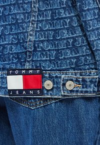 Prednji dio traper jakne s plavim tkaninom na kojoj se ponavlja tekst branda, ukrašen pravokutnim logotipom u crvenoj, bijeloj i mornarsko plavoj boji. Srebrni gumbi.