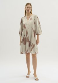 Beige klänning med kort ärm och abstrakta rosa mönster, V-ringning, sammanknuten detalj vid axlarna, svävande silhuett, i kombination med silverfärgade sandaler.
