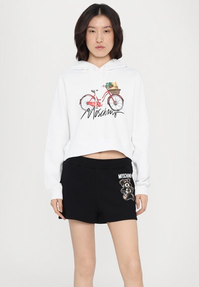 Balta trumpa pūkinė striukė su raudonu dviračio grafiku ir tekstu "Moschino"; derinama su juodais šortais, kuriuose yra meškos dizainas ir logotipas.