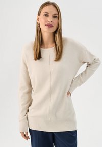 Beżowy sweter dziany z okrągłym dekoltem, długimi rękawami, prążkowanymi mankietami i prążkowanym dołem. Posiada centralny szew jako dodatkowy element wzorniczy.