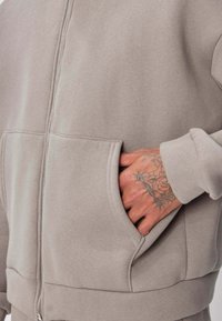 Main avec des tatouages floraux et d'étoiles partiellement insérée dans la poche avant d'un sweat à capuche beige à fermeture éclair.