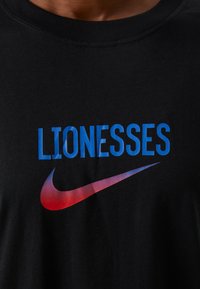 Camiseta negra con el texto azul "LIONESSES" y un logo de swoosh de Nike en degradado rojo. Diseño de cuello redondo, material de algodón, textura suave.