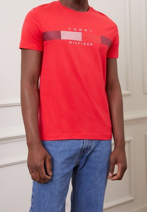Homme portant un t-shirt rouge Tommy Hilfiger avec un logo rayé sur la poitrine et un jean bleu, debout à l'intérieur.