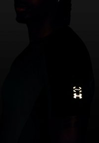 Mann, der ein dunkles Shirt trägt, mit einem leuchtenden "Halo" und dem Under Armour-Logo auf dem oberen Arm im schwachen Licht.