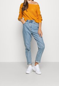 Pull orange épaules dénudées en maille épaisse, associé à un jean taille haute bleu clair et des baskets blanches, avec des revers roulés et une coupe décontractée.