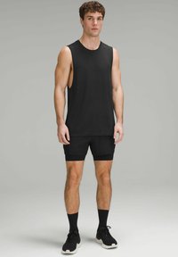Sort ærmeløs atletisk tanktop med et tekstureret strikstof, parret med sorte shorts. Model iført sorte ankelsokker og sneakers.