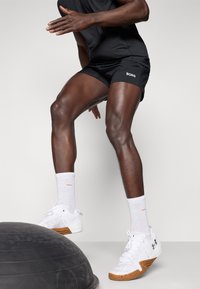 Zwarte sportshorts met "BORG" logo, witte sportschoenen met textuurdetails en witte sokken met oranje accenten.