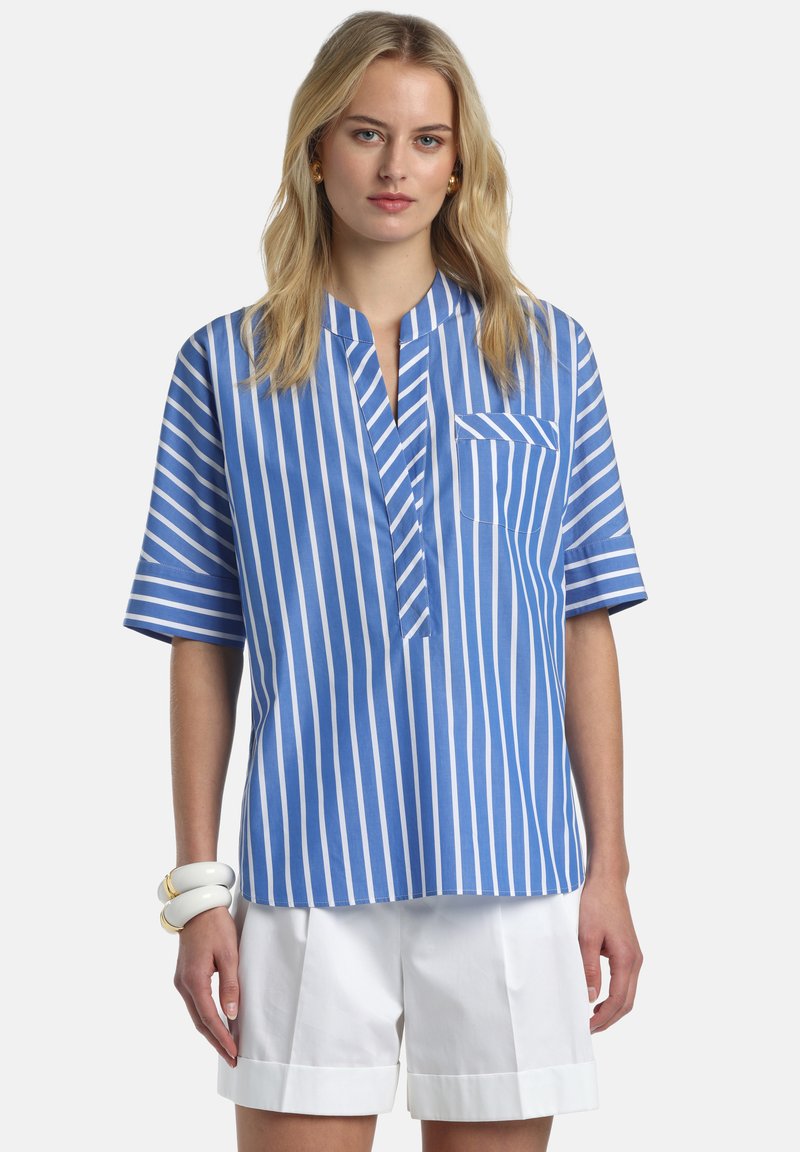 Jeune femme portant une chemise à manches courtes à rayures verticales bleues et blanches et un short blanc à ourlet roulé, avec des boucles d'oreilles dorées et des bracelets blancs.