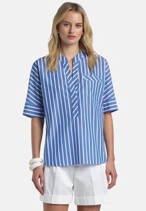 Young woman wearing a blue and white vertical striped short-sleeve shirt and white rolled-hem shorts, with gold earrings and white bracelets.