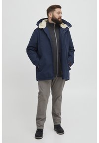 Marineblå jakke med faux fur-fôr på hetta, glidelås lukking og knappedetaljer på lommene, sammen med grå bukser og svarte sko.