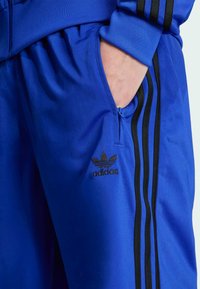 Kék sportnadrág sima textúrával, cipzáras zsebbel, fekete Adidas logóval és három fekete csíkkal az oldalán.