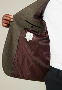 Blazer marrón texturizado con un forro estampado en burdeos, que cuenta con un bolsillo en el pecho y una etiqueta visible en su interior. Diseño de ajuste regular.