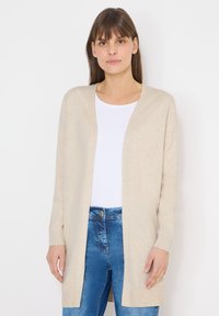 Femme portant un cardigan beige ouvert sur un haut blanc et un jean bleu, debout devant un fond blanc uni.