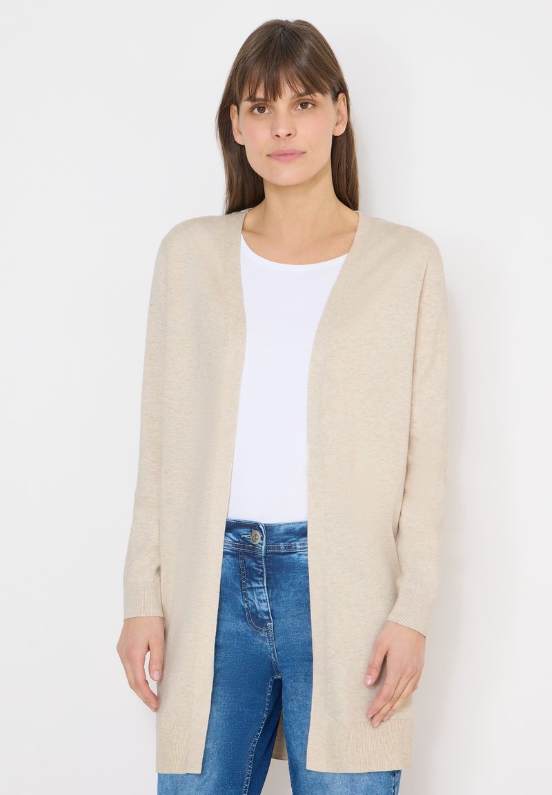 Femme portant un cardigan beige ouvert sur un haut blanc et un jean bleu, debout devant un fond blanc uni.