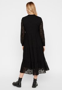 Kvinna klädd i en lång svart spetsklänning med genomskinliga ärmar och svarta ankelboots, stående vänd bort från kameran mot en neutral vit bakgrund.