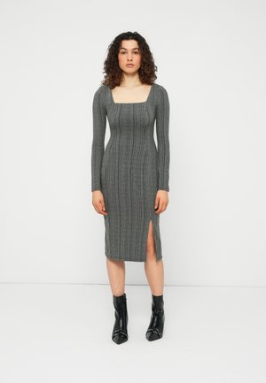 Femeie cu păr creț, purtând o rochie gri închis, tricotată, cu mâneci lungi şi o despicătură frontală, asortată cu ghete negre până la gleznă.