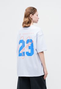 Biele oversized tričko s nápisom "THE GREATEST" v ružovej farbe, "23" v modrej farbe a "Jordan" v oranžovej kurzívou na chrbte.