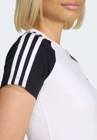 Biele tričko s čiernymi krátkymi rukávmi. Na ramene má tri biele pruhy. Hladká bavlnená textúra, dizajn s okrúhlym výstrihom.