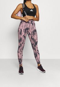 Czarny sportowy top z krótkim rękawem z białym logo, w połączeniu z różowo-czarnymi legginsami w wzory. Model nosi czarne buty sportowe.