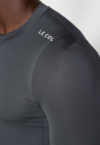 Maillot de cyclisme gris foncé avec un tissu texturé, design ajusté, et logo "LE COL" sur l'épaule. Matériau lisse et respirant.