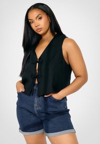 Svart kortärmad väst med djup V-ringning och framknutar, i kombination med blå denimshorts med uppvikta fållar och sidfickor.