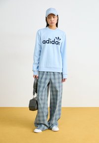 Sudadera Adidas de color azul claro con logo negro, combinada con pantalones de pierna ancha de cuadros. Los accesorios incluyen una gorra azul clara y un bolso negro pequeño.