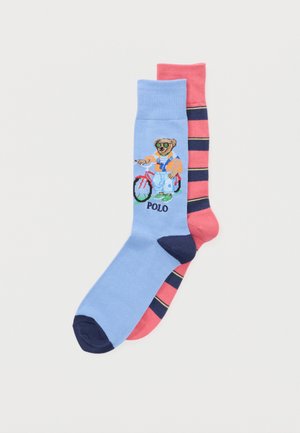 Deux chaussettes colorées : une bleue avec un ours portant des lunettes de soleil et une salopette à côté d'un vélo avec le texte "POLO", l'autre rose avec des rayures bleu marine et jaunes.