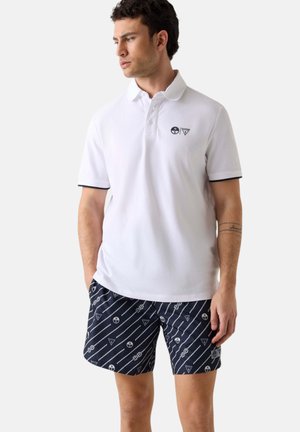 Junger Mann trägt ein weißes Poloshirt mit dunklem Besatz und marineblaue Shorts mit diagonalen Streifen und nautischen Motiven.