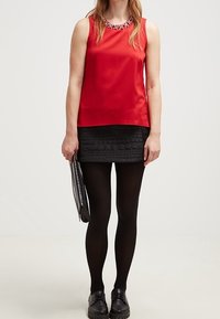 Top rosso senza maniche con scollatura decorata di gioielli, abbinato a una mini gonna nera con texture. La modella indossa collant neri e scarpe, tenendo una pochette argento.