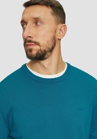 Teal gebreide trui met een ronde hals en een witte binnenkraag. Bevat subtiele branding op de borst. Effen en textielstof.