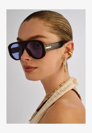 Nő, aki hátrafésült hajjal, nagy fekete napszemüveget, arany karika fülbevalókat, rétegezett gyöngyös nyakláncokat és krémszínű kötött felsőt visel.