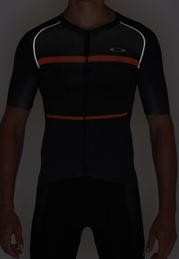 Musta, istuva pyöräilypaita lyhyillä hihoilla, oransseilla vaakasuorilla raidoilla ja valkoisilla yksityiskohdilla. Varustettu etuvetoketjulla ja teksturoidulla kangasmallilla.