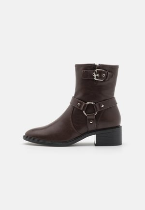 Bottine marron en simili cuir avec zip latéral, détail boucle et petit talon carré. Présente une texture lisse et un design à bout arrondi.
