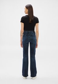 Sieviete ar gariem tumšiem matiem, valkājot melnu īsrokainu topu, tumši zilas zvana bikses, baltas kurpes un tetovējumus uz kreisās rokas, stāvot ar muguru uz priekšu.