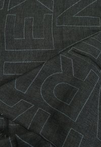 Tessuto in denim scuro con grandi contorni blu chiaro di lettere. La trama appare liscia con una leggera lucentezza. Il denim è piegato.