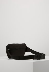 Marsupio in tessuto nero di forma rettangolare, con chiusura a zip e cinturino regolabile in tessuto. Presenta una texture liscia e un design minimale.
