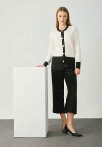 Cardigã creme com detalhes pretos e grandes botões, combinado com calças largas pretas e sapatos de salto pontiagudo pretos. Textura de tecido suave.