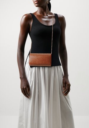 Femme portant un haut noir sans manches, une jupe plissée blanche, des boucles d'oreilles en or, un collier et un petit sac bandoulière marron avec une chaîne.