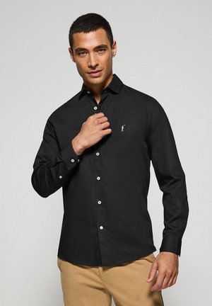 Camisa negra de manga larga con cuello, detalles de botones blancos y un pequeño logo en el pecho. Textura suave y ajuste entallado.