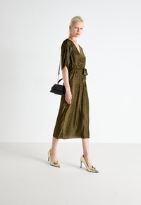 Metallic olive-green dress with a wrap design, short sleeves, and waist tie. Paired with gold high-heeled shoes and a black handbag.