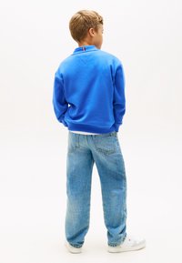 Μπλε βαμβακερό φούτερ με γιακά και κόκκινη και λευκή ριγέ λεπτομέρεια στο πίσω μέρος. Συνδυασμένο με ανοιχτό μπλε τζιν παντελόνι και λευκά αθλητικά παπούτσια.