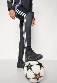 Zwarte sportbroek met witte strepen, zwarte voetbalschoenen, en een voetbal met een zwart-wit sterrenpatroon, met neonaccenten.