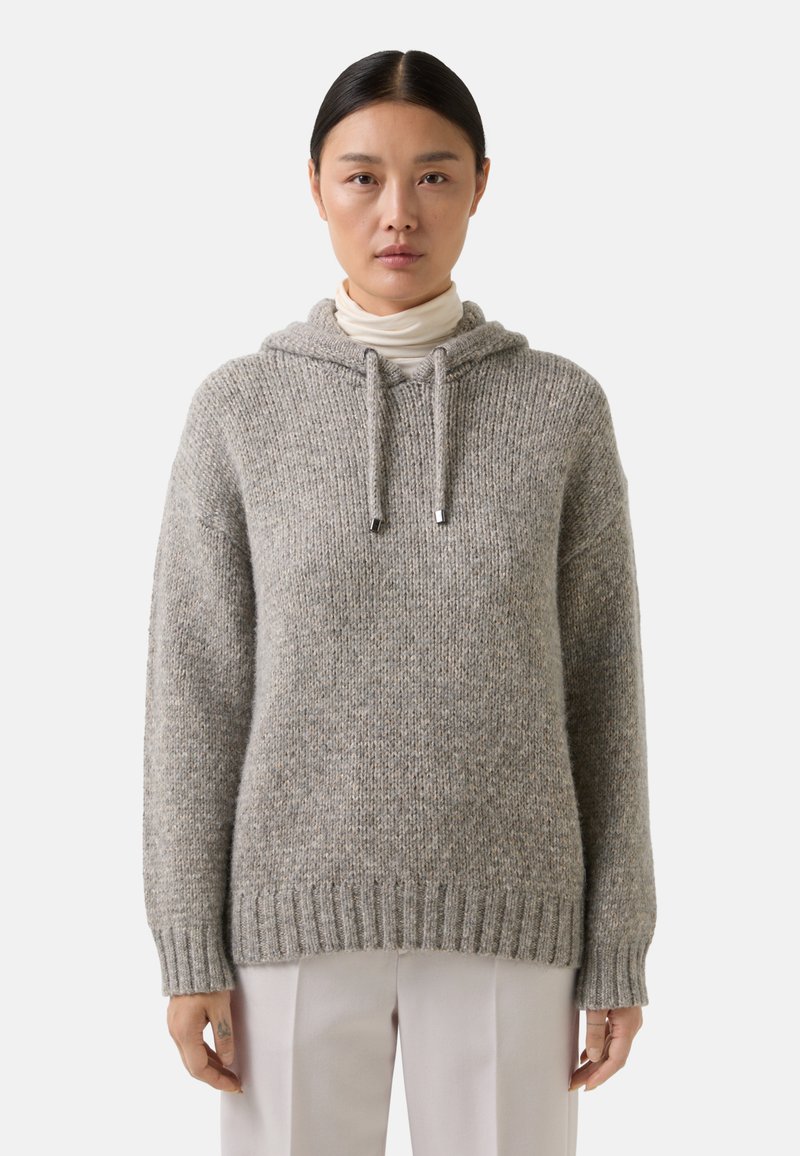 Vrouw met stijl zwart haar die een grijze gebreide hoodie-sweater over een witte coltrui en lichtgekleurde broek draagt, staande.
