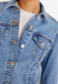 Veste en denim bleu clair avec un col classique, des boutons pression en métal argenté et deux poches poitrine. Présente des coutures orange et une coupe décontractée.