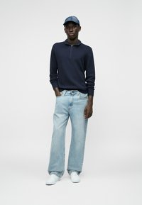 Polo lungo a maniche con zip in tessuto blu navy, abbinato a jeans in denim a gamba larga di colore azzurro e sneaker bianche, completato da un cappellino in denim.