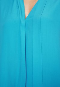 Gros plan sur un vêtement turquoise avec un col en V et un détail plissé sur le devant.